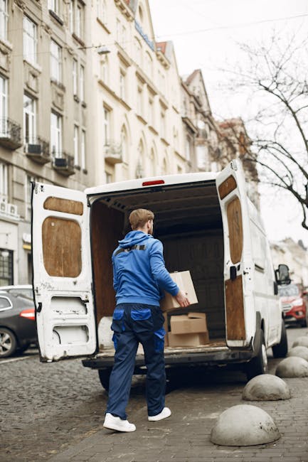 A man with short blond hair, dressed in a bright blue hoodie and dark blue trousers, is seen loading a cardboard box into the open rear of a white cargo van parked on a cobbled street in Putney. The van, which is part of a house removals service by Man with Van Putney, has its rear doors wide open, revealing the interior lined with wooden panels and a black cargo floor. Inside the van, several other boxes of various sizes, some wrapped in plastic or packing paper, are visible. The man is standing on the pavement, which is separated from the street by rounded stone bollards, and appears to be carefully positioning or lifting a box in preparation for home relocation or furniture transport. In the background, multi-story Victorian-style buildings with ornate facades, balconies, and large windows line the street, along with trees that have shed their leaves for the season. Additional parked cars are visible along the roadside, emphasizing the urban environment, and the scene is lit by natural daylight, indicating a clear day suitable for packing and moving activities.