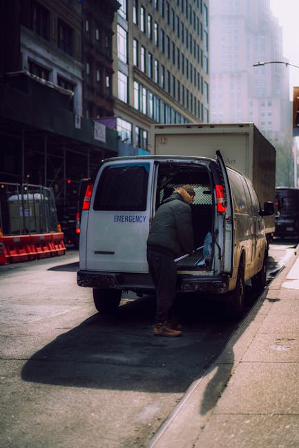 A man with short blond hair, dressed in a bright blue hoodie and dark blue trousers, is seen loading a cardboard box into the open rear of a white cargo van parked on a cobbled street in Putney. The van, which is part of a house removals service by Man with Van Putney, has its rear doors wide open, revealing the interior lined with wooden panels and a black cargo floor. Inside the van, several other boxes of various sizes, some wrapped in plastic or packing paper, are visible. The man is standing on the pavement, which is separated from the street by rounded stone bollards, and appears to be carefully positioning or lifting a box in preparation for home relocation or furniture transport. In the background, multi-story Victorian-style buildings with ornate facades, balconies, and large windows line the street, along with trees that have shed their leaves for the season. Additional parked cars are visible along the roadside, emphasizing the urban environment, and the scene is lit by natural daylight, indicating a clear day suitable for packing and moving activities.