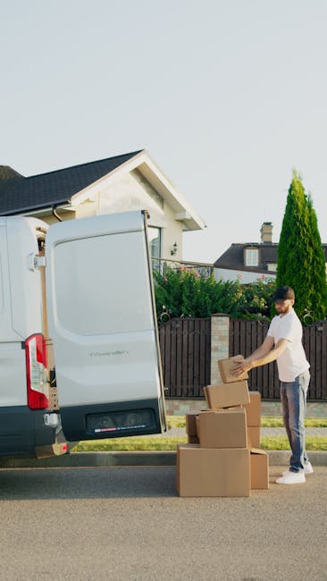 A white moving van parked on a residential street in Putney, with the side door open and two men involved in a house relocation process. One man is seated inside the van, possibly the driver, wearing