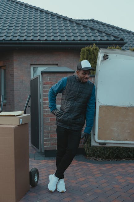 A man dressed in a blue long-sleeve shirt, black padded vest, black trousers, and white footwear stands outdoors near a white van with a partially open side door, engaged in home relocation activities. In the background, there is a brick house with a tiled roof, a small planting hedge, and a paved driveway. The man is leaning against the van, appearing to rest during a loading process involving furniture or household items, as evidenced by large cardboard boxes and a hand trolley on the pavement nearby. The scene is set during daytime with natural light, and the environment suggests a neighbourhood setting typical for house removals, with visible packaging materials and equipment used for furniture transport and packing during a move. This setup aligns with professional removals services like those offered by Man with Van Putney, supporting efficient packing, loading, and home relocation logistics.
