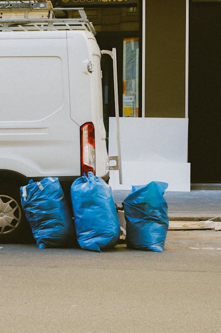 Close-up view of the rear section of a white commercial van parked on a street in Putney, with the back doors closed. In front of the van, there are three large blue plastic bags filled with bulky waste, possibly furniture or household items, placed on the pavement. The bags appear to be securely tied, indicating they are ready for removal or disposal. The van is positioned near a building with a glass door and windows, suggesting an entrance to a property or commercial premises. The area is during daylight, with natural light illuminating the scene, capturing the loading process involved in home relocation or decluttering activities. Man with Van Putney's services likely include such furniture transport and waste removal, helping clients manage bulky items during moves or property clearances, with a focus on efficient, same-day pickup options.
