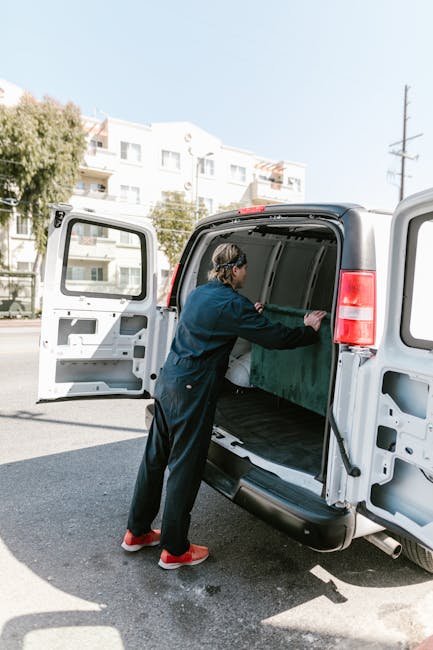 A man dressed in a blue long-sleeve shirt, black padded vest, black trousers, and white footwear stands outdoors near a white van with a partially open side door, engaged in home relocation activities. In the background, there is a brick house with a tiled roof, a small planting hedge, and a paved driveway. The man is leaning against the van, appearing to rest during a loading process involving furniture or household items, as evidenced by large cardboard boxes and a hand trolley on the pavement nearby. The scene is set during daytime with natural light, and the environment suggests a neighbourhood setting typical for house removals, with visible packaging materials and equipment used for furniture transport and packing during a move. This setup aligns with professional removals services like those offered by Man with Van Putney, supporting efficient packing, loading, and home relocation logistics.