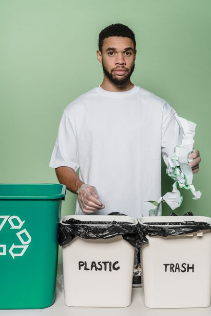 Inside a room with a light blue wall, clear plastic containers labeled 'PAPER' and 'PLASTIC' with green recycling symbols are positioned on a flat surface, containing waste materials. The 'PAPER' cont