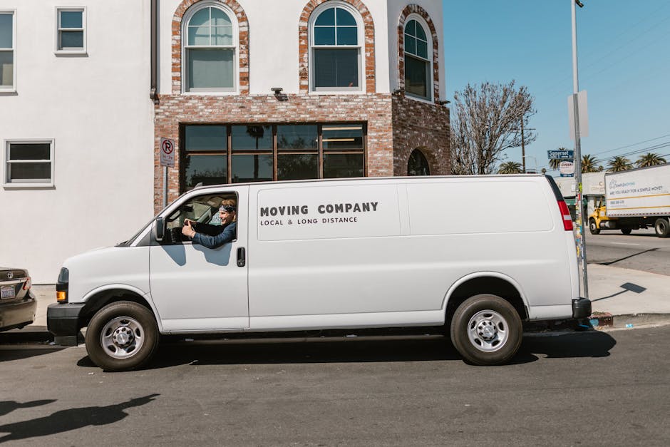 A white moving van from Man with Van Putney, used for house and flat relocation services, parked parallel to a curb on a city street in front of a brick building with large glass windows. The van feat