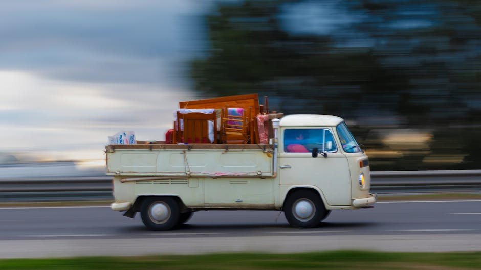 A white moving van from Man with Van Putney, used for house and flat relocation services, parked parallel to a curb on a city street in front of a brick building with large glass windows. The van feat