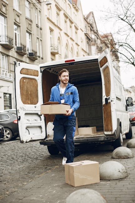 A man with a beard wearing a blue jacket and navy work trousers is standing at the back of a white moving van, carrying a medium-sized cardboard box. The van’s rear doors are open, revealing a spacious interior with more packed boxes and packing materials. Several other cardboard boxes are placed on the cobbled pavement next to the van, suggesting an ongoing home relocation process. The scene is set outside on a city street with classic multi-storey residential buildings in the background, featuring large windows and balconies. The pavement has a few large rounded stones, and the overall environment is overcast, indicating typical moving day conditions. This image illustrates furniture transport and packing steps involved during a professional house removal, as provided by Man with Van Putney, aligning with services such as removals and furniture transport for a move from Putney High Street to SW15.