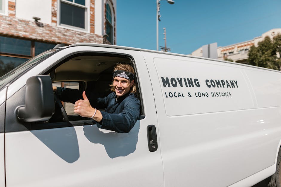 A smiling male mover with long hair, wearing a black bandana and a dark blue shirt, is sitting in the driver's seat of a white delivery van during a home relocation process on Putney's narrow streets.