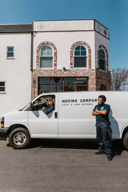 A white moving van parked on the street in front of a two-storey residential building with a white and brick exterior, featuring arched windows on the upper floor. The van is labeled with the words 'Moving Company, Local & Long Distance' on the side. Inside the driver's cabin, a person with light-colored hair and sunglasses is engaged in conversation with a second individual standing beside the van. The person outside, wearing a dark jacket and standing with arms crossed, appears to be a mover involved in home relocation or furniture transport. The scene is set during daylight with clear weather, and nearby trees are visible in the background. The image captures elements relevant to packing, loading, and moving logistics associated with house removals, in alignment with the services offered by Man with Van Putney.