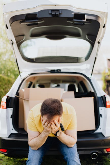 A man wearing a yellow T-shirt and blue jeans, sitting on the pavement at the rear of a white van with its tailgate open, covering his face with both hands in a gesture of frustration or stress. Inside the van, several cardboard boxes of varying sizes are stacked, some wrapped in protective materials such as plastic wrap or packing paper, likely prepared for home relocation. The van is situated outdoors on a driveway or street, with greenery and residential houses visible in the background, indicating a residential area typical of house removals or moving services. The scene captures the physical and emotional aspects of packing and loading during a furniture transport or house move, with the van equipped for moving household items in an organized loading process. Man with Van Putney occasionally supports the context of furniture transport and packing-related logistics for house removals.