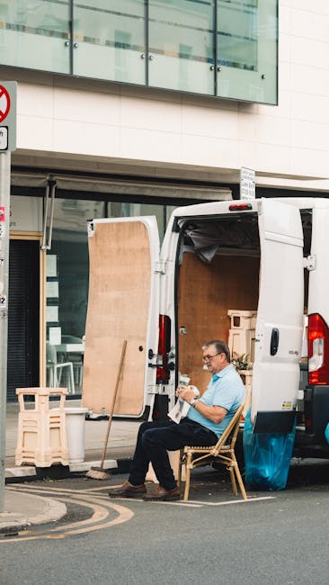 A man with glasses wearing a light blue shirt and dark trousers is seated on a wooden chair beside the open rear doors of a white moving van parked on a city street. The van's interior is visible, showing wooden panels and empty cardboard boxes, indicating an ongoing home relocation or furniture transport process. In front of the van, there are wooden pallets and a small folding table. The scene takes place in front of a modern building with large glass windows and a white facade. The man appears to be taking a break during packing and loading activities, which are part of house removal services provided by Man with Van Putney. The pavement is visible, with a no-parking sign nearby, and the lighting suggests daytime conditions. The setting reflects a typical urban moving scenario involving packing materials, loading equipment, and transportation logistics.