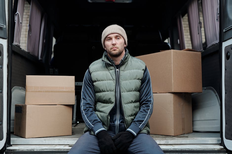 A young man with a neutral facial expression, wearing a grey knit beanie, a green and navy puffer vest over a navy jacket, black gloves, and dark trousers, sits inside the open back of a moving van on a paved surface. Behind him, the van's interior is visible, showing several cardboard boxes of various sizes, some sealed with packing tape, placed on the floor and leaning against the van's sidewalls, which are lined with protective lining. The boxes are packed in an organized manner, prepared for home relocation or furniture transport. The loading area appears well-lit with natural light. The scene is a still image capturing a moment in the packing and loading process associated with professional removals or moving services, emphasizing furniture and packing materials, as well as the vehicle used for transport, in a neutral and factual manner. Man with Van Putney's logo or branding is not visible in the image, maintaining a focus on the objects and person present.