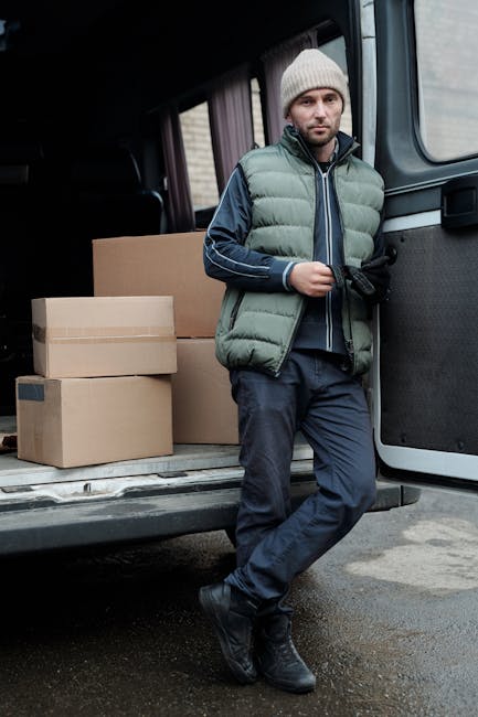 A man with glasses wearing a light blue shirt and dark trousers is seated on a wooden chair beside the open rear doors of a white moving van parked on a city street. The van's interior is visible, showing wooden panels and empty cardboard boxes, indicating an ongoing home relocation or furniture transport process. In front of the van, there are wooden pallets and a small folding table. The scene takes place in front of a modern building with large glass windows and a white facade. The man appears to be taking a break during packing and loading activities, which are part of house removal services provided by Man with Van Putney. The pavement is visible, with a no-parking sign nearby, and the lighting suggests daytime conditions. The setting reflects a typical urban moving scenario involving packing materials, loading equipment, and transportation logistics.