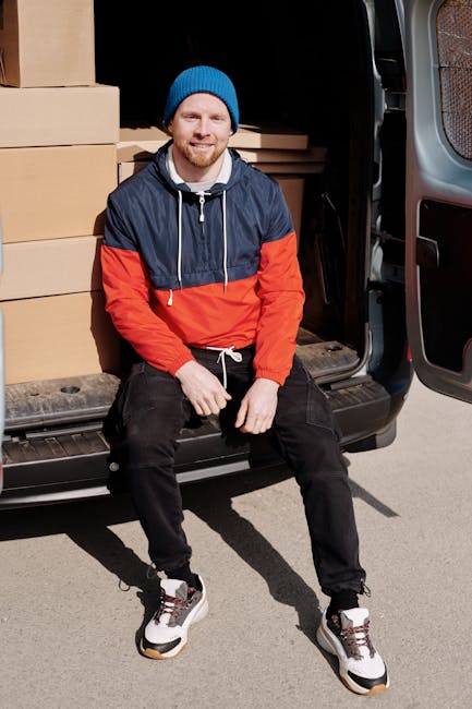 A man with a beard, wearing a blue beanie, a navy and red waterproof jacket, black trousers with white drawstrings, and sneakers, is sitting on the edge of an open black van parked on a driveway. The van's interior is visible behind him, filled with neatly stacked cardboard boxes and packing materials, including plastic wraps and brown paper. The boxes vary in size and are arranged on the vehicle’s floor and side shelves, indicating a home relocation process involving furniture and items being prepared for transport. The man appears relaxed, possibly taking a break during loading or unloading, with his hand resting on his knee. The surrounding area is well-lit, suggesting daytime, and the context is related to professional removals or furniture transport, supported by the presence of packing materials and the open vehicle ready for moving activities. This scene aligns with house removals, packing, and loading procedures associated with home relocation services offered by Man with Van Putney, as referenced on their website page about Wandsworth Council skip rules.