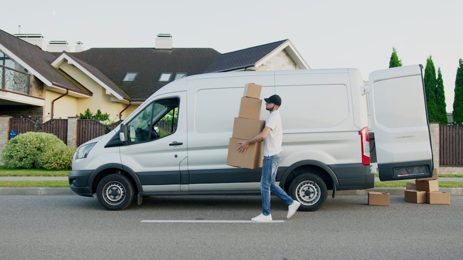 A man wearing a white T-shirt, jeans, white sneakers, and a black cap is loading four large cardboard boxes into the back of a white cargo van parked on a residential street. The van is positioned parallel to the pavement in front of a row of modern, semi-detached houses with pitched roofs, some featuring skylights. The vehicle's side door is closed, while the rear doors are open, revealing the empty interior where the boxes are being placed. Additional cardboard boxes are on the pavement beside the van, and a trolley or hand truck might be involved in the loading process. The scene is outdoors during daylight with natural lighting, and a neatly maintained garden with bushes and tall, narrow trees lines the front yards of the houses, indicating a suburban setting suitable for home relocation, furniture transport, and packing and moving services provided by Man with Van Putney.