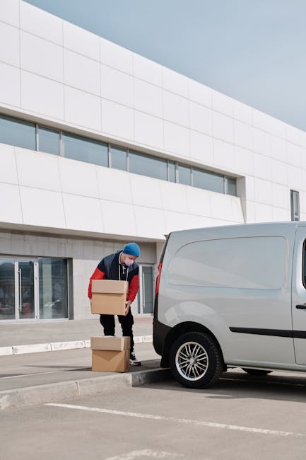 A man with a beard, wearing a blue beanie, a navy and red waterproof jacket, black trousers with white drawstrings, and sneakers, is sitting on the edge of an open black van parked on a driveway. The van's interior is visible behind him, filled with neatly stacked cardboard boxes and packing materials, including plastic wraps and brown paper. The boxes vary in size and are arranged on the vehicle’s floor and side shelves, indicating a home relocation process involving furniture and items being prepared for transport. The man appears relaxed, possibly taking a break during loading or unloading, with his hand resting on his knee. The surrounding area is well-lit, suggesting daytime, and the context is related to professional removals or furniture transport, supported by the presence of packing materials and the open vehicle ready for moving activities. This scene aligns with house removals, packing, and loading procedures associated with home relocation services offered by Man with Van Putney, as referenced on their website page about Wandsworth Council skip rules.